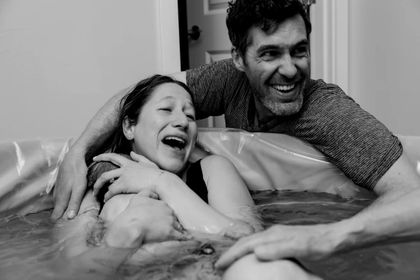 A father wraps his arms around his partner as she holds their newborn baby in the birth tub after their home birth, which was partially covered by Medicaid