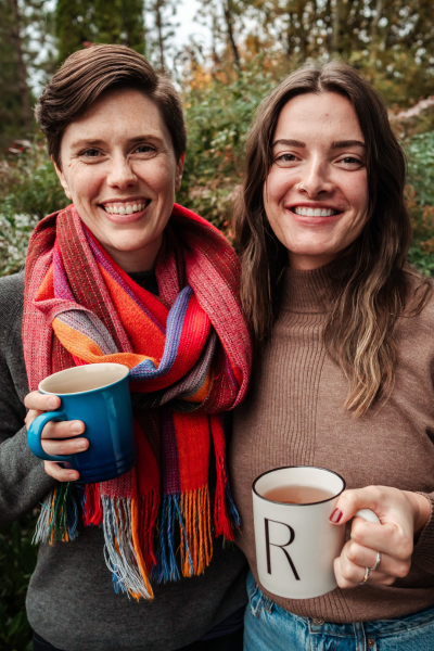 Puget Sound midwives Caroline Sausler (she/her), LM, CPM and Rachel Wright (she/her) CPM, MSM, BSN have their arms around one another as they smile into the camera holding mugs
