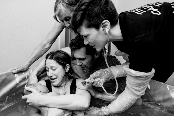 Black and white photo of a Puget Sound mother in a birth tub holding her newborn baby with her husband, mother, and midwife by her side