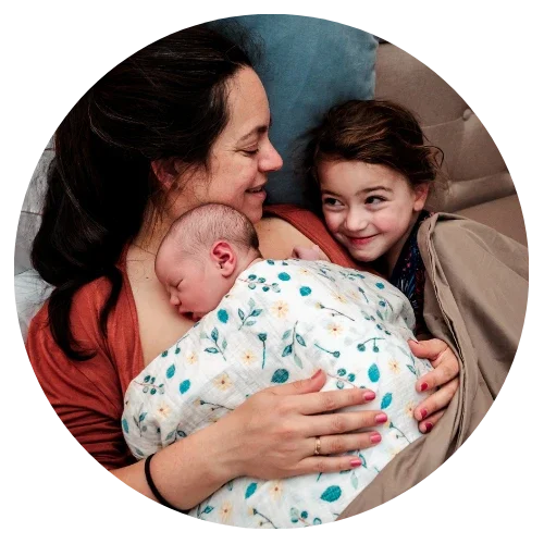A mother cuddles in bed with her newborn baby and older daughter after having a home birth with Wildflower Midwifery and Fertility